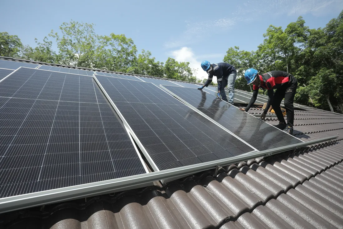 Installers carrying a solar panel across a rooftop in Langkawi, Kedah, surrounded by dense greenery