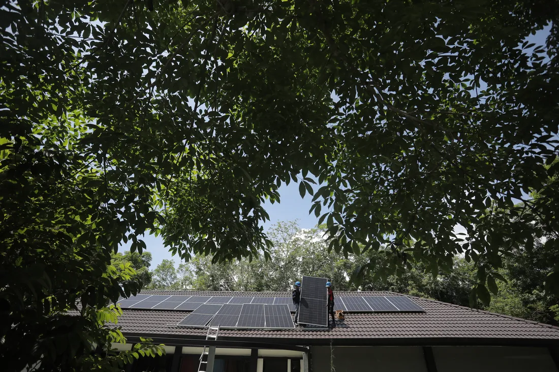 Two installers lifting a solar panel into place on a pitched roof in Langkawi against open sky